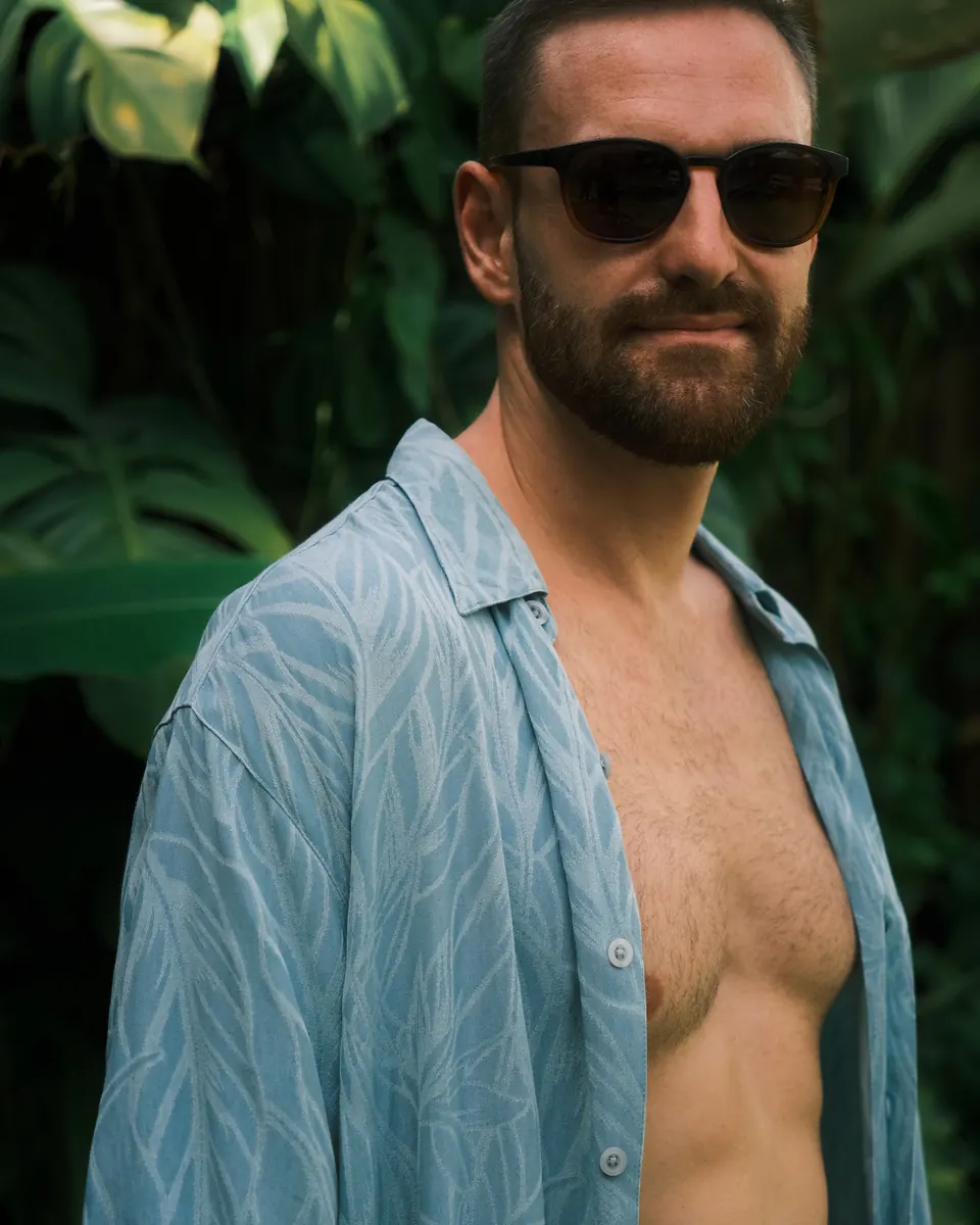 Porträt von Timon Kukwa mit Sonnenbrille und offenem Hemd vor tropischen Pflanzen