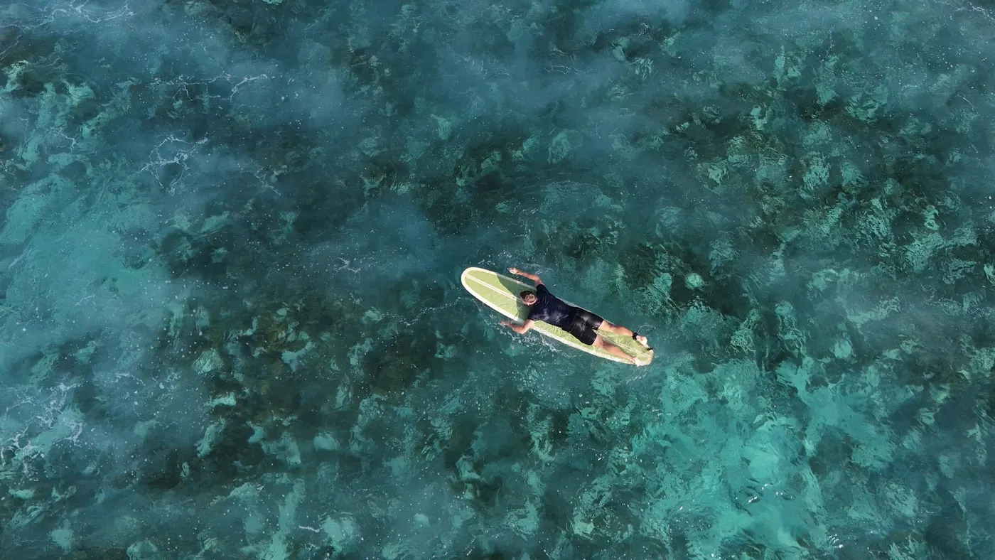 Drohnenaufnahme: Timon Kukwa paddelt auf seinem Surfbrett über türkisfarbenes Wasser und ein Riff