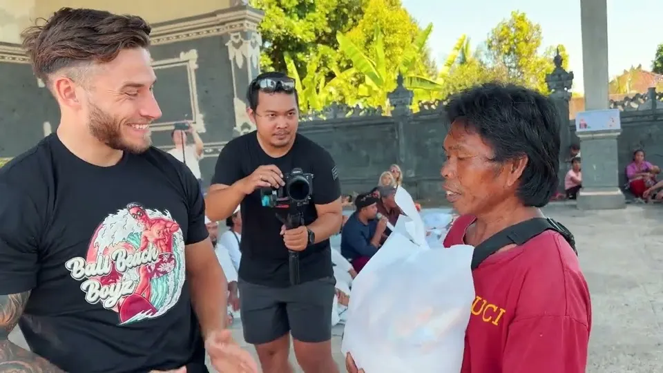 Ein Helfer übergibt einer Frau vor einem Tempel eine Tasche mit Lebensmitteln, ein Kameramann filmt