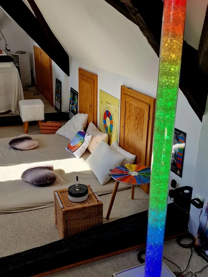 Eine Lichtsäule in Regenbogenfarben in einem hellen Raum mit Sitzkissen und Bildern an der Wand