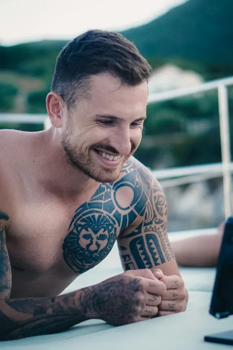 Nico Hartmann mit Tattoos auf Schulter und Armen, lachend vor seinem Smartphone