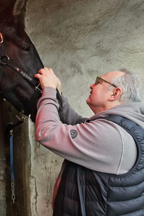 Thorsten Bach in Weste und Kapuzenjacke legt einem Pferd im Stall die Hände an den Kopf und lächelt es an